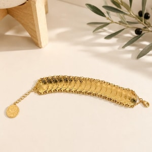 May include: A gold-toned bracelet composed of a series of small, circular, coin-like discs linked together in a row. The bracelet features a delicate chain extension at one end, finished with a single dangling coin charm and a lobster claw clasp.