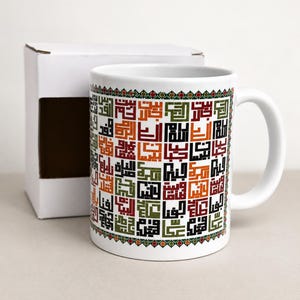 May include: White ceramic mug featuring a colorful geometric pattern of Arabic calligraphy. The design includes squares of text in shades of red, green, and orange, with a decorative border. A white box is next to the mug.