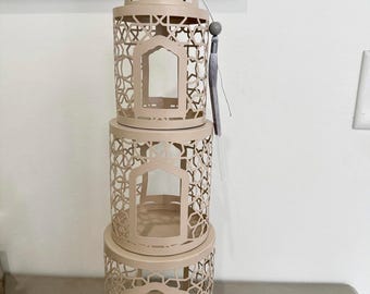 May include: A stacked set of three beige metal lanterns with intricate geometric cutouts. Each lantern has a rectangular window and a circular base. The top lantern is topped with a gold crescent moon and star finial. A gray tassel hangs from the top.