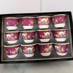 May include: A set of twelve small, decorative coffee cups. The cups are a deep magenta with a white base and feature gold accents, including crescent moons, stars, and a mosque design. The cups are arranged in a black box.