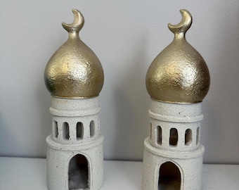 May include: Two decorative ceramic mosque-shaped candle holders. Each has a gold-colored dome with a crescent moon finial and a white, speckled base with arched openings. The candle holders are designed to hold candles.