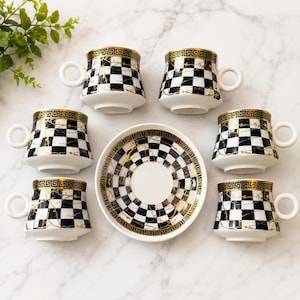May include: A set of six ceramic coffee cups and one matching saucer featuring a black and white checkered pattern with gold marble-like veining and a gold Greek key border along the top rim. Each cup has a round handle and a white interior base.