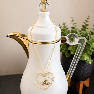 May include: A white and gold teapot with a heart-shaped pendant. The pendant is clear with gold flakes and the Arabic word "Shai". The teapot has a gold handle and accents, and a clear glass top.