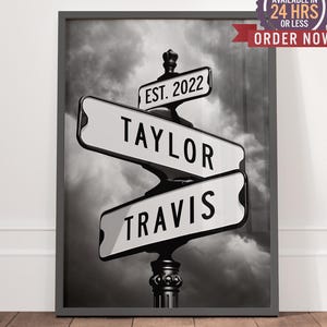 May include: Black and white framed print featuring a street sign design. The sign displays the names "Taylor" and "Travis" with "Est. 2022" above. The background is a cloudy sky. The print includes a "Preview Available in 24 Hrs or Less" badge.