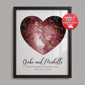 May include: A framed wall art print featuring a heart-shaped dark red nebula and star constellation map. The text reads: Gabe and Michelle, Forever began beneath these stars, December 12, 2025. The print is enclosed in a simple black frame.