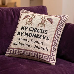 以下が含まれることがあります： 「MY CIRCUS MY MONKEYS」の文字とAnna、Daniel、Katherine、Josephの名前が書かれた正方形の装飾用クッション。クッションにはボーダーデザインと猿のイラストが描かれています。紫色の豪華なソファの上に置かれています。