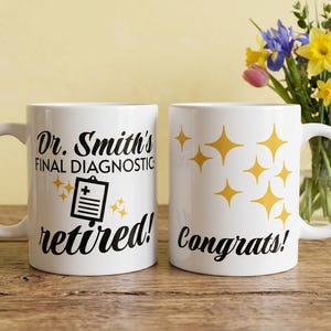 Può includere: Due tazze di ceramica bianca. Una tazza dice "Dr. Smith's Final Diagnostic retired!" con una grafica di cartella clinica. L'altra tazza dice "Congrats!" con stelle dorate. Un mazzo di fiori è sullo sfondo.