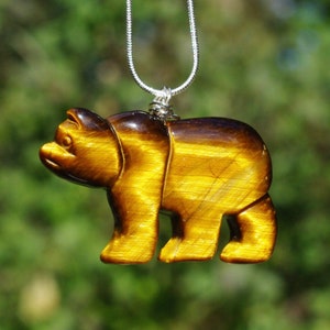 May include: A pendant necklace featuring a carved bear pendant made of tiger's eye stone. The bear is a warm golden brown color with darker brown accents. The necklace chain is a thin, silver-colored metal.