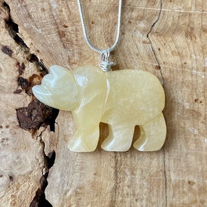 May include: A yellow carved bear pendant on a silver chain. The pendant is made of a translucent stone with a light yellow color.