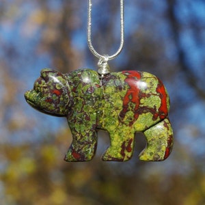 May include: A green and red bear pendant carved from a stone with a marbled pattern. The bear is hanging from a silver chain.