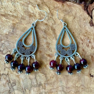 May include: Silver-tone teardrop earrings with a filigree design and dangling dark red beads. The earrings have a hook closure and are displayed on a wooden surface. The earrings are a bohemian style.