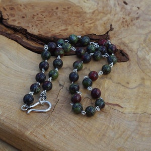May include: A green and red gemstone beaded necklace with a silver clasp. The necklace is made of round beads and is approximately 20 inches long.