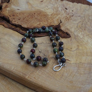 May include: A green and red gemstone beaded necklace with a silver clasp. The necklace is on a wooden surface.