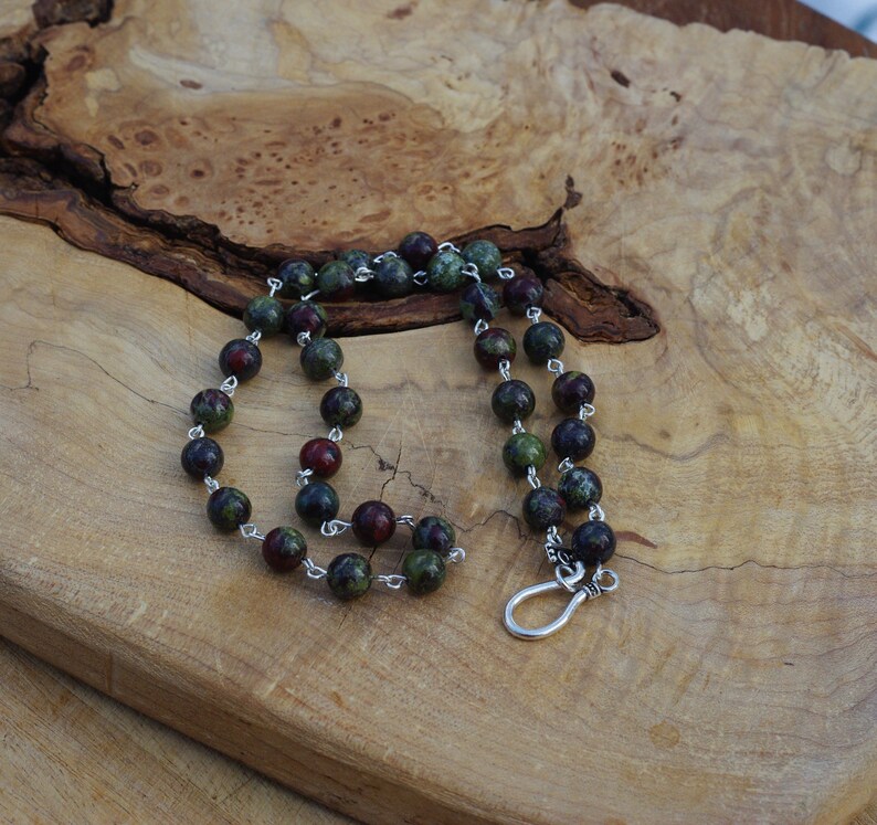 May include: A green and red gemstone beaded necklace with a silver clasp. The necklace is on a wooden surface.