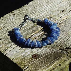 Op de afbeelding: Een blauwe edelsteen armband met zilveren kettingschakels. De armband is gemaakt van kleine, platte, blauwe stenen die in een rij aan elkaar geregen zijn. De armband ligt op een houten oppervlak.
