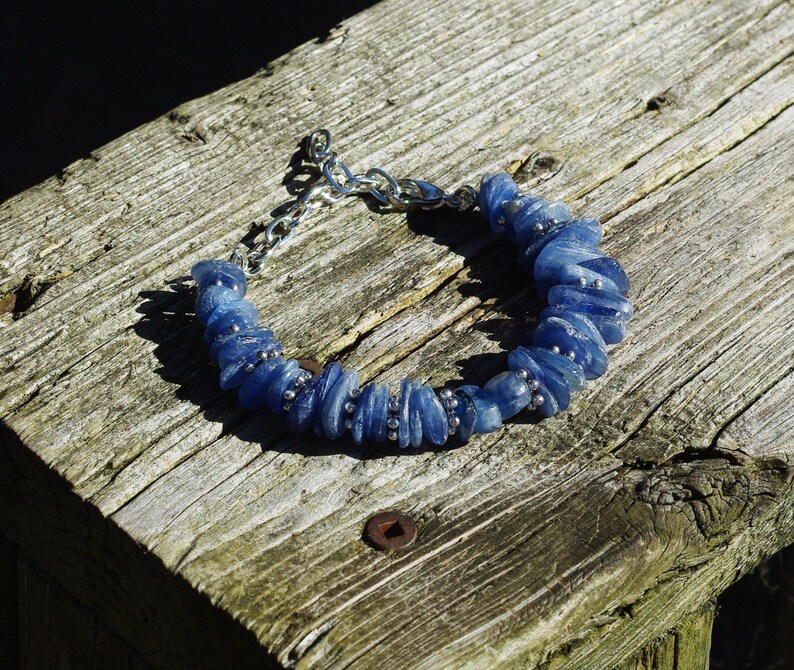 Op de afbeelding: Een blauwe edelsteen armband met zilveren kettingschakels. De armband is gemaakt van kleine, platte, blauwe stenen die in een rij aan elkaar geregen zijn. De armband ligt op een houten oppervlak.