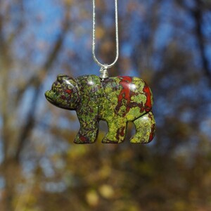 May include: A green, red, and brown carved bear pendant on a silver chain. The bear is a detailed carving with a mottled pattern.
