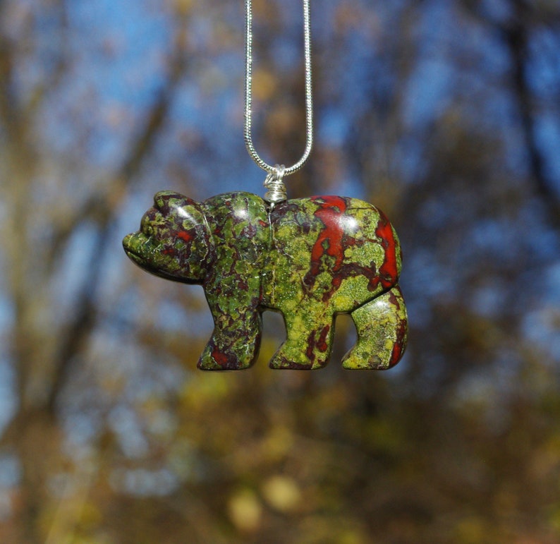 May include: A green, red, and brown carved bear pendant on a silver chain. The bear is a detailed carving with a mottled pattern.