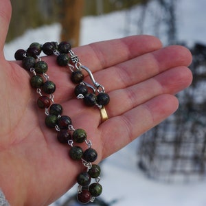 May include: A green and red gemstone beaded necklace with a silver clasp. The necklace is on a hand.