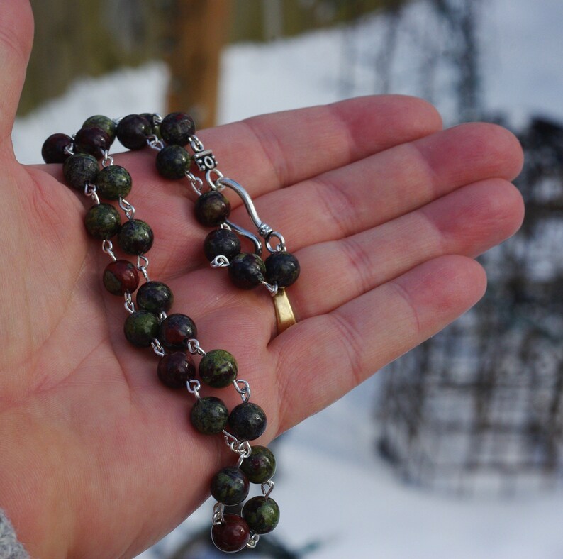 May include: A green and red gemstone beaded necklace with a silver clasp. The necklace is on a hand.