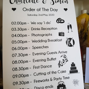 Könnte beinhalten: Ein weißes Schild mit schwarzem Text und Symbolen, die die Tagesordnung für eine Hochzeit auflisten. Die Hochzeit ist für Charlotte und Simon am Samstag, den 2. Mai 2020. Der Zeitplan umfasst die Zeremonie, den Empfang, das Abendessen, das Tanzen und das Feuerwerk.