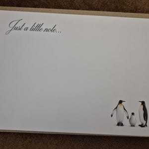 May include: A white stationery card with the text "Just a little note..." in black script. The card features a penguin family illustration at the bottom. The card is set inside a brown envelope.