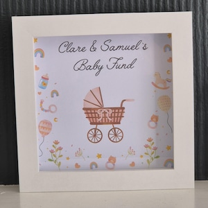 May include: A framed print with the text "Clare & Samuel's Baby Fund" above a brown baby carriage illustration. The print is decorated with baby-themed illustrations in pastel colors, including rainbows, balloons, and flowers.