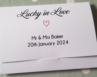 Puede incluir: Una tarjeta blanca con el texto "Lucky in Love" en escritura negra, un corazón rosa y los nombres "Mr & Mrs Baker" con la fecha "20 de enero de 2024" impresos en negro. La tarjeta está doblada.