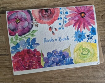 May include: A watercolour floral greeting card with the text "Thanks a Bunch" in blue script. The card features flowers in purple, red, pink, blue, and yellow. The card is on a brown envelope, set against a wooden background.
