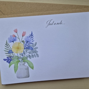 Puede incluir: Una tarjeta de papelería blanca con un diseño floral en acuarela y el texto "Just a note...". El arreglo floral presenta flores amarillas, azules y rosas en un jarrón gris. La tarjeta está sobre un sobre marrón.