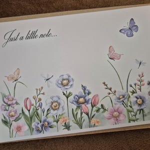 May include: A floral greeting card with the text "Just a little note...". The card features watercolour illustrations of flowers, butterflies, and dragonflies in soft pastel colours. The card is set against a neutral background.
