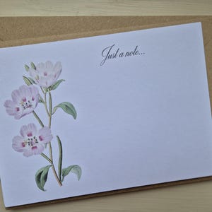 Könnte beinhalten: Eine weiße Schreibwarenkarte mit einer floralen Illustration von rosa Blüten und grünen Blättern, mit dem Text "Just a note..." in schwarzer Schrift. Die Karte liegt auf einem braunen Umschlag.