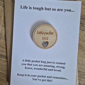 Può includere: Un piccolo abbraccio tascabile in legno con un cuore, su un biglietto bianco all'interno di una busta marrone. Il biglietto ha un testo: "La vita è dura, ma anche tu..." e "Ce la puoi fare!" Il disco di legno dice "piccolo HUG tascabile".