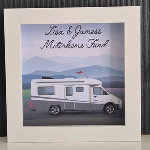 May include: A framed print featuring a white and gray motorhome with a red flag, set against a backdrop of layered blue mountains. The text "Lisa & James's Motorhome Fund" is written above the vehicle.