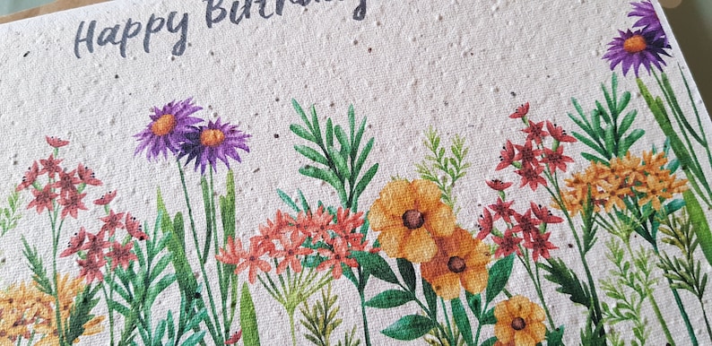Może przedstawiać: Biała kartka z tekstem "Happy Birthday" napisanym czarnym atramentem. Kartka jest ozdobiona akwarelową ilustracją r&oacute;żnych kwiat&oacute;w w odcieniach fioletu, ż&oacute;łtego, pomarańczowego i czerwonego.