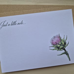May include: A white card with the handwritten text "Just a little note...". The card features a detailed illustration of a purple thistle flower with green leaves. It is placed on a brown envelope.