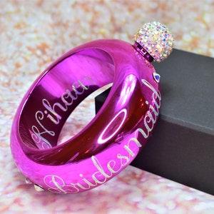 May include: A pink metal bracelet with a crystal-encrusted top. The bracelet is engraved with the words "Bridesmaid" and "[Name]" in a script font. The bracelet comes with a silver funnel.