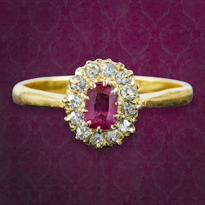 Pode incluir: Um anel de ouro com uma pedra preciosa central de corte retangular em rosa profundo, rodeada por um halo de doze pequenas pedras redondas e claras facetadas. O aro é de ouro liso e polido, criando um design clássico de agrupamento.