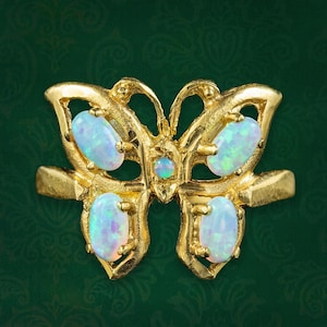 May include: A gold ring featuring a butterfly-shaped centerpiece. The butterfly design is accented with four oval-shaped iridescent opals on the wings and one small round opal in the center, all set within a polished gold metal frame.