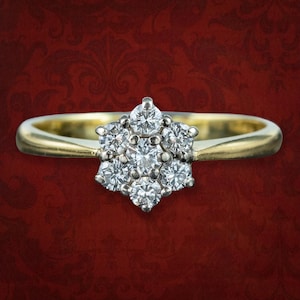 May include: A gold ring with a cluster of diamonds in a floral arrangement. The ring's band is a polished gold, and the diamonds are set in a silver-colored metal. The ring is set against a red background.