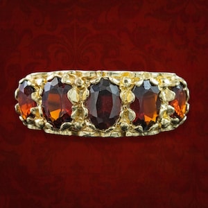 Puede incluir: Un anillo de oro con cinco piedras preciosas de granate rojo oscuro, talladas en forma ovalada y dispuestas en una fila horizontal. La banda de metal presenta un diseño texturizado y orgánico con pequeños detalles dorados en relieve entre las piedras.