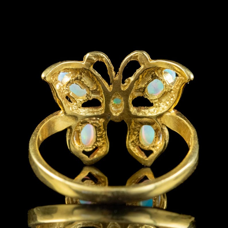 May include: A gold ring featuring a butterfly-shaped centerpiece adorned with five small, iridescent oval-shaped opals. The metal band is smooth and circular, supporting the detailed butterfly motif which displays a textured, openwork design.