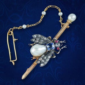 May include: A gold and silver brooch in the shape of a bee with a pearl body, blue sapphire, and red ruby accents. The brooch is attached to a gold chain with pearls.