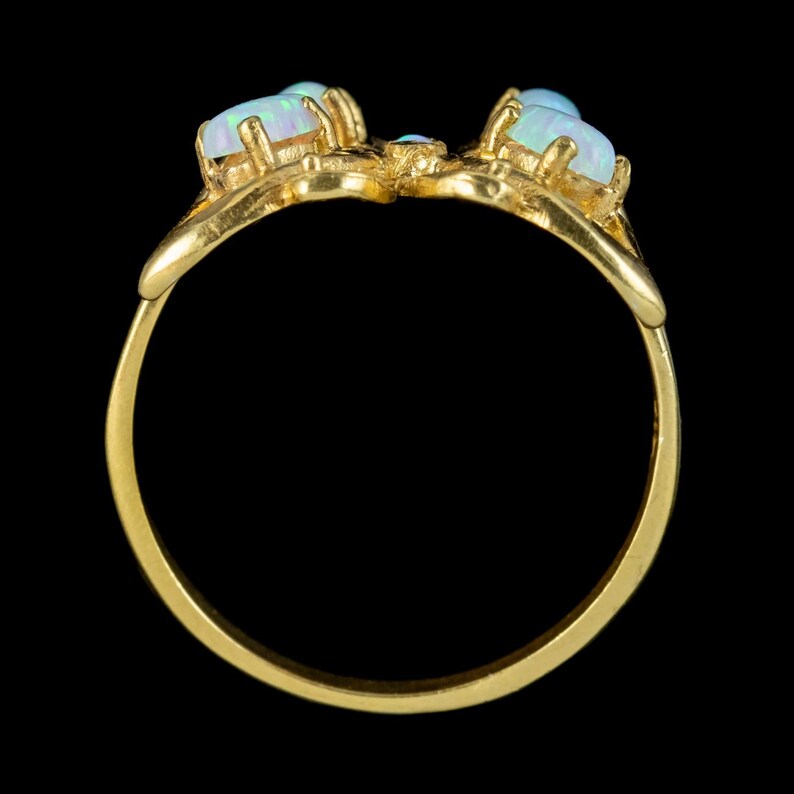 May include: A gold ring featuring a band with a decorative top section set with three cabochon opals. The opals display iridescent flashes of blue and green light, held in place by gold prongs. The ring has a smooth, polished gold finish.