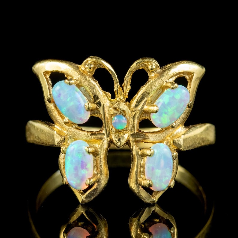 May include: A gold ring featuring a butterfly-shaped centerpiece. The butterfly design is accented with four oval-shaped iridescent opals on the wings and one small round opal in the center of the body, all set within a polished gold metal frame.