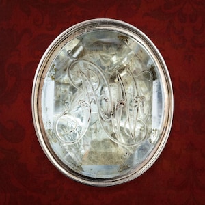 May include: An antique, oval-shaped silver compact mirror with an engraved monogram. The mirror's surface is slightly aged, showing signs of wear. The background is a deep red with a subtle, ornate pattern.