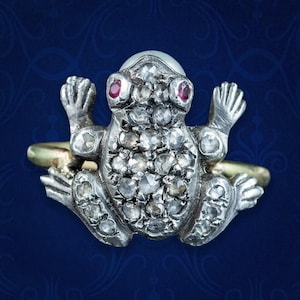 May include: A silver frog ring with ruby eyes and a pearl on its head. The frog's body is encrusted with small, sparkling diamonds. The ring band is gold. The ring is set against a dark blue background.