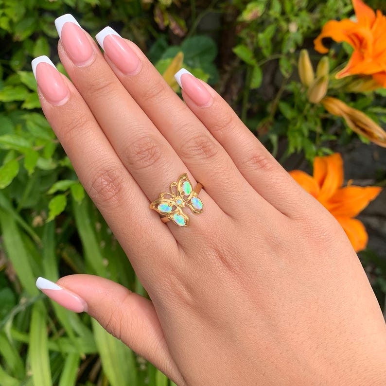 May include: A gold-toned ring featuring a butterfly-shaped centerpiece with four iridescent, oval-shaped opal-like stones set into the wings. The metal band is thin and polished, showcasing a delicate openwork design on the butterfly wings.