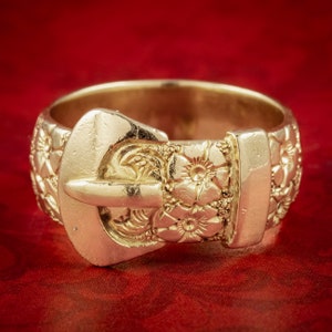 May include: A gold-toned ring with a detailed buckle design. The ring is decorated with floral patterns and has a smooth, polished finish.
