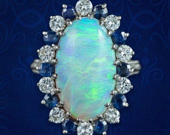 May include: An oval opal ring with a halo of diamonds and sapphires. The large opal has a pearlescent, iridescent quality. The diamonds and sapphires are set in a silver or white gold band, creating a striking contrast against the opal's colors.